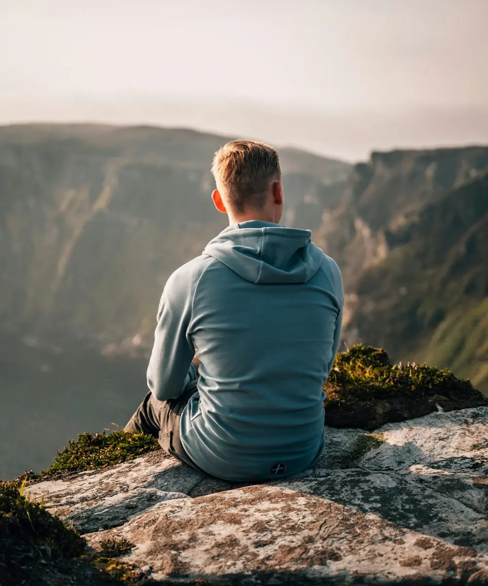 A man sitting quietly on a cliff edge, looking out over a green valley